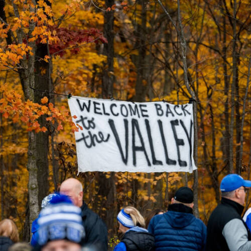 Sign reads Welcome Back to the Valley, colorful fall trees in background, people gathering in winter hats and coats